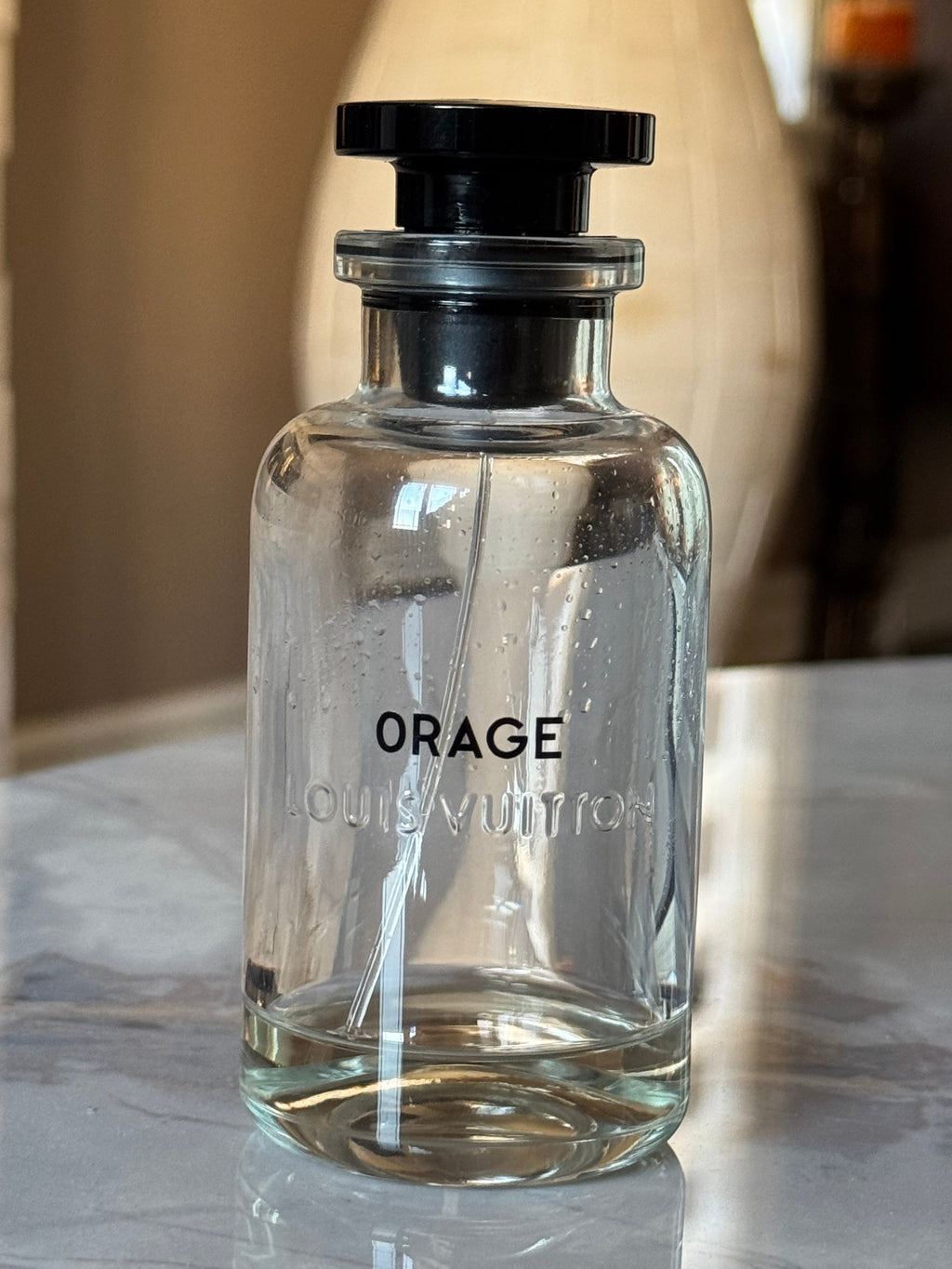 A clear glass perfume bottle with a black cap, featuring the text 'ORAGE' and 'LOUIS VUITTON' embossed on the front, sitting on a marble countertop.